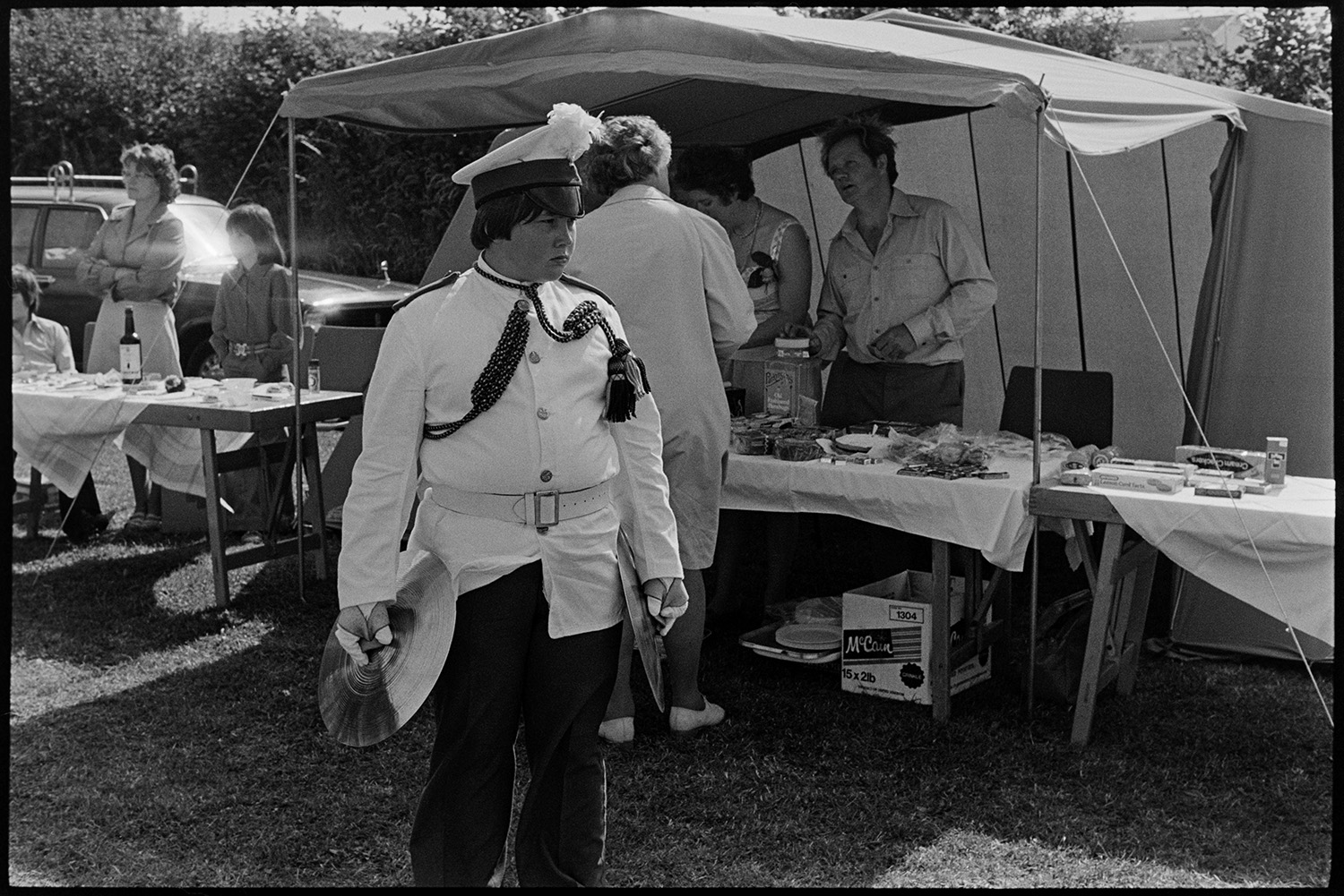 Band and drum majorettes on parade. 
[A band member holding cymbals at Atherington Village fete. He is stood by a stall selling sweets.]
