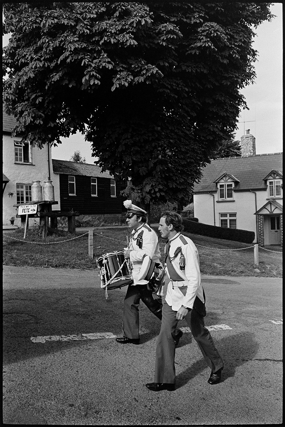 Bandsman walking back after playing at fete. 
[Two bandsmen walking home after performing at Atherington Village fete. One of them is carrying a drum. Two milk churns are on a stand under a tree in the background.]
