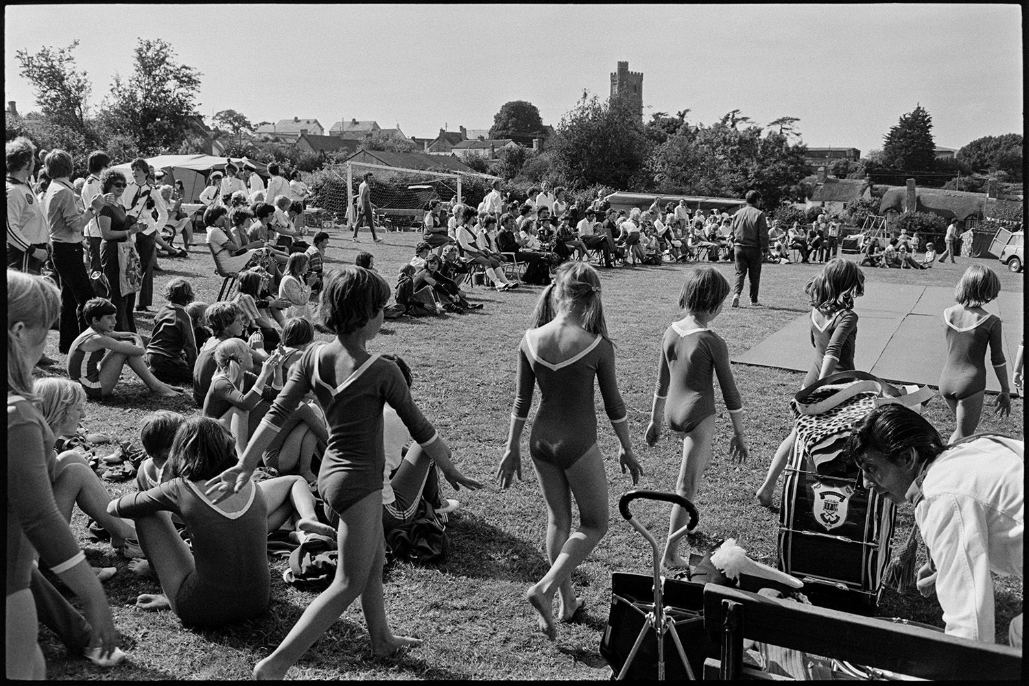 Gymnasts at fete. 
[Girl gymnasts walking out to perform at Atherington Village fete. A crowd has gathered to watch them.]