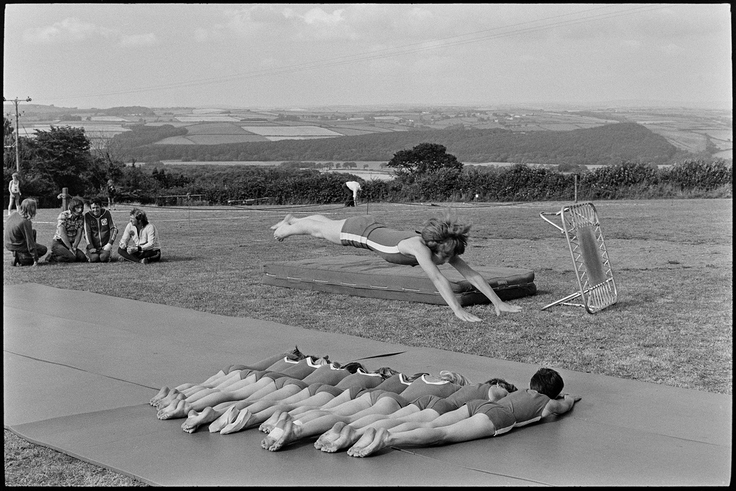 Gymnasts and fancy dress at fete. 
[Gymnasts performing a routine in a field at Atherington fete. One gymnast is jumping over nine other gymnasts lying on the ground. People are watching the routine in the background.]