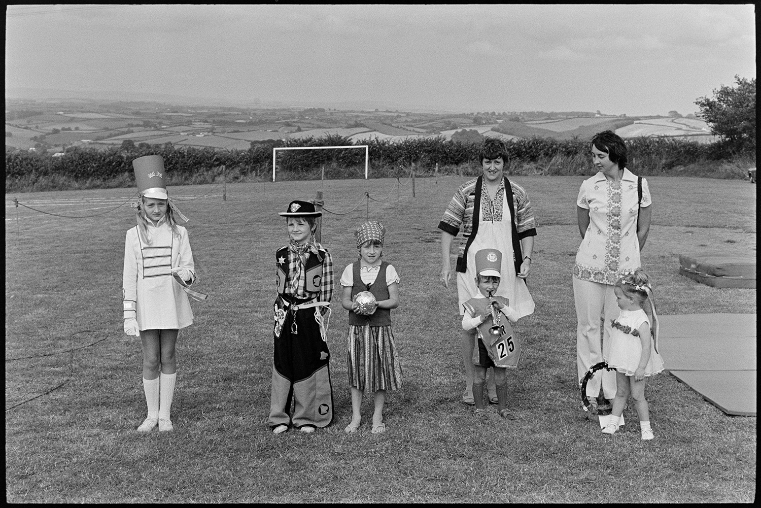 Gymnasts and fancy dress at fete. 
[Five children in fancy dress in a field at Atherington fete. Costumes include a soldier, cowboy and fortune teller. A landscape of fields can be seen in the background.]
