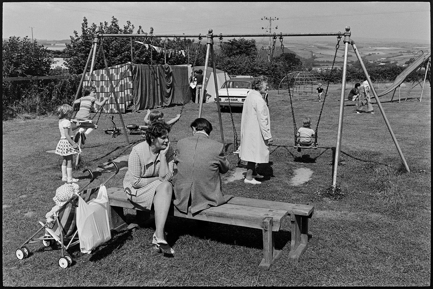 Families, swings, pony rides at village fete. 
[Children playing on a slide, climbing frame and swings in a field at Atherington fete. A man and woman are sat on a bench by a pushchair and another woman is pushing a child on a swing. Tents and a parked car are visible in the background.]