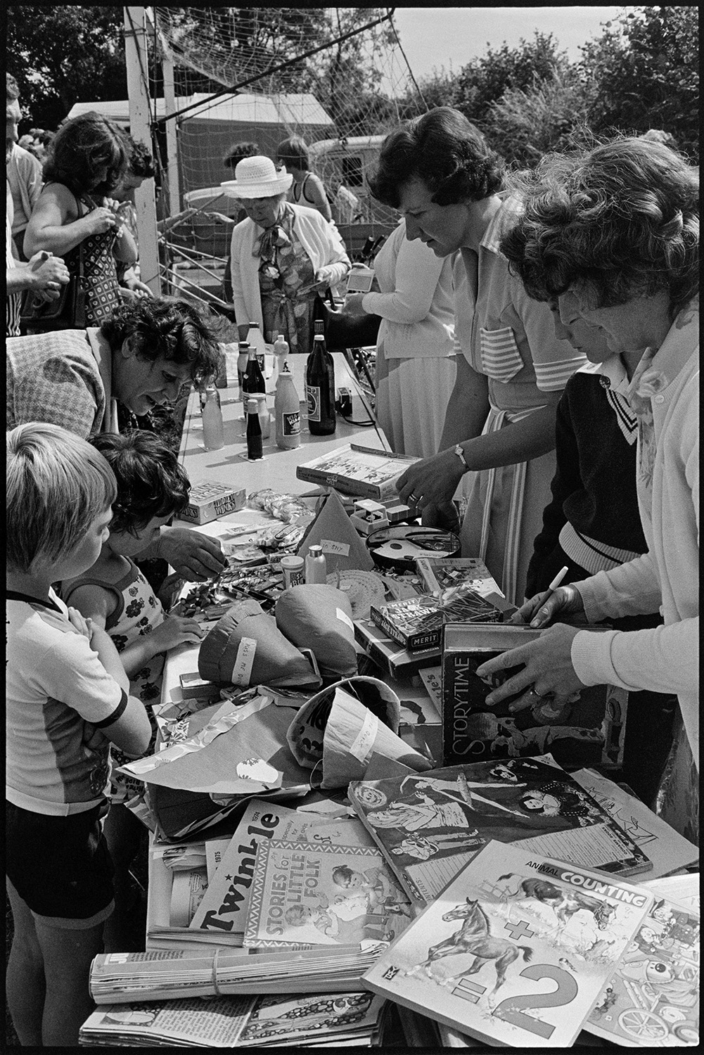 People buying from stall at village fete. 
[Women and children looking at items on a stall at Atherington fete. Items on the stall include books and games.]