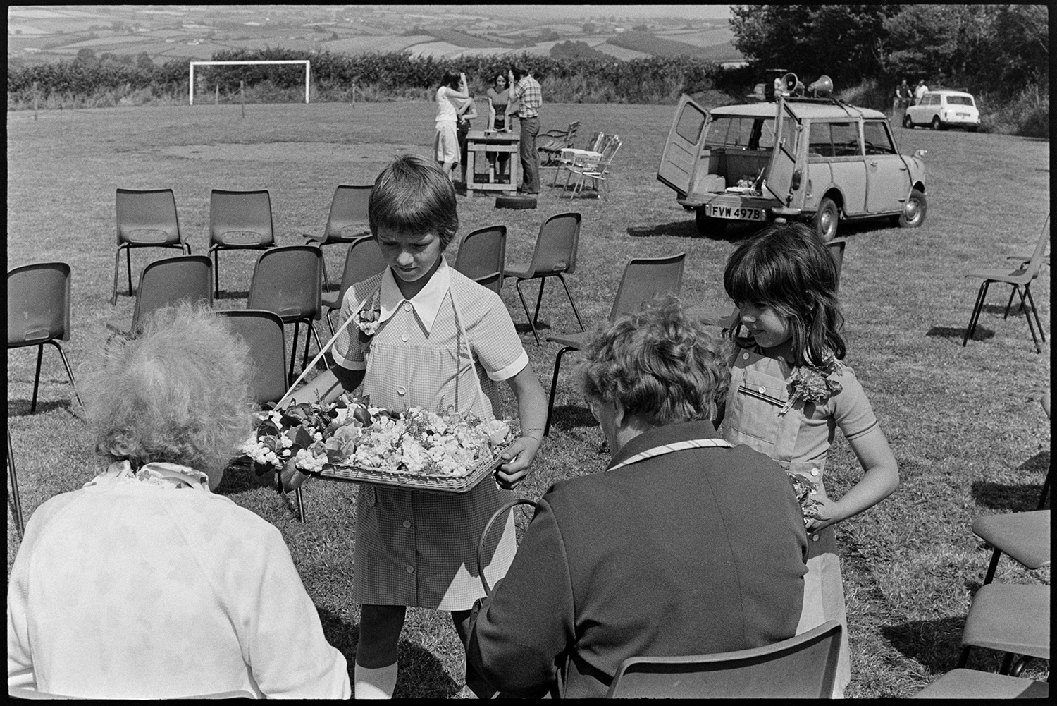 Children selling flowers at village fete. 
[Two girls selling flowers to two women at Atherington fete. A van with loudspeakers is parked in the field in the background.]