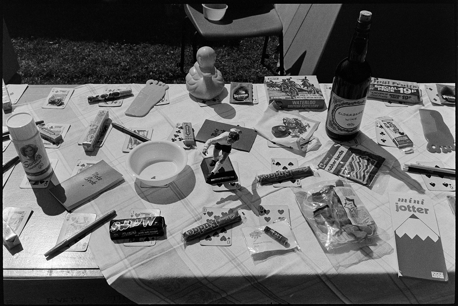 Items for sale, stall at fete. 
[Prizes which can be won on a game at Atherington fete. Each prize is on a playing card. Prizes include chocolate bars, notebooks, wine and figurines.]
