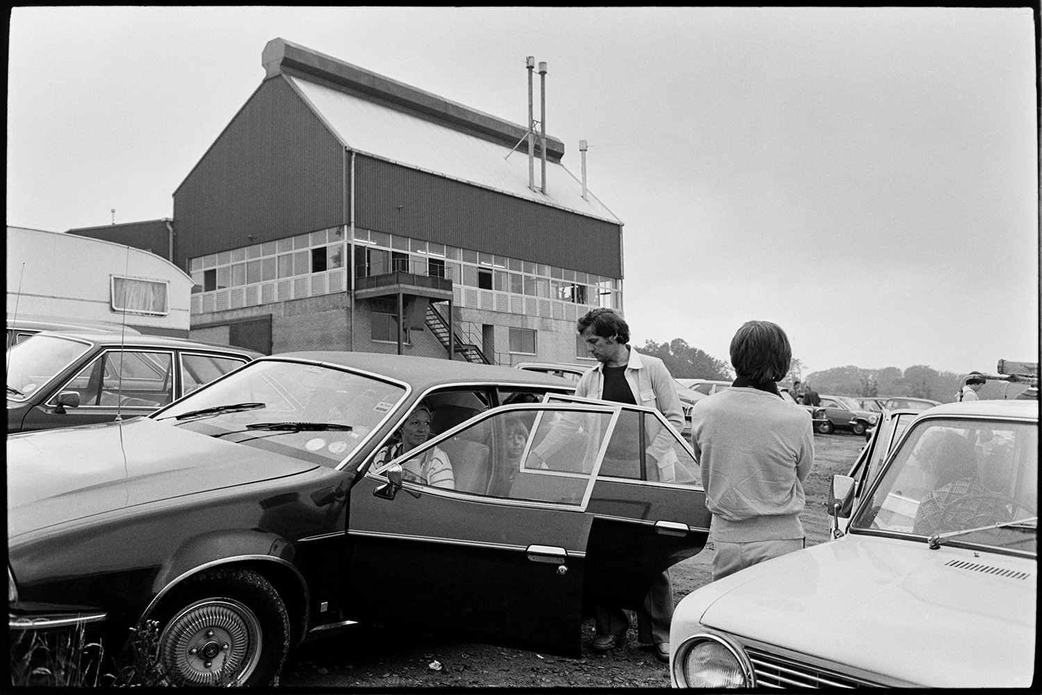 Management and workers at glass factory. 
[Visitors to the Dartington Glass factory, also known as Dartington Crystal, in Torrington, by their parked car in the car park. The factory building is visible in the background.]
