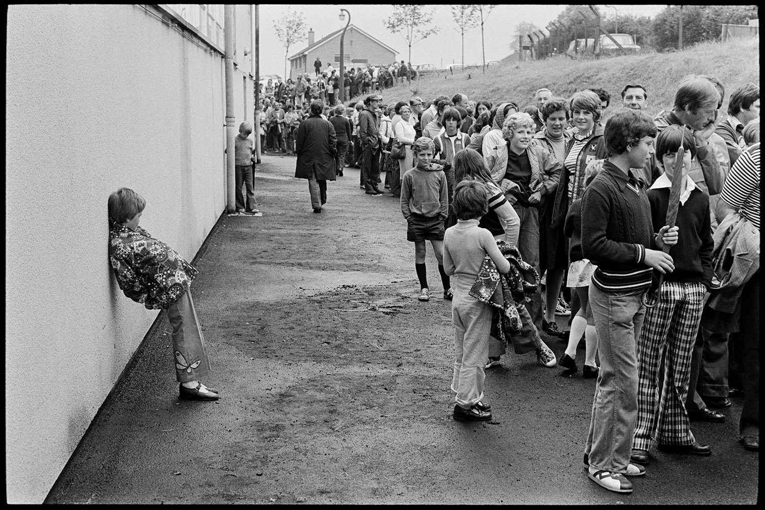 Management and workers at glass factory. 
[People queuing up to visit the Dartington Glass factory, now known as Dartington Crystal, at Torrington. Men, women and children are queuing and one child is leaning against a wall.]