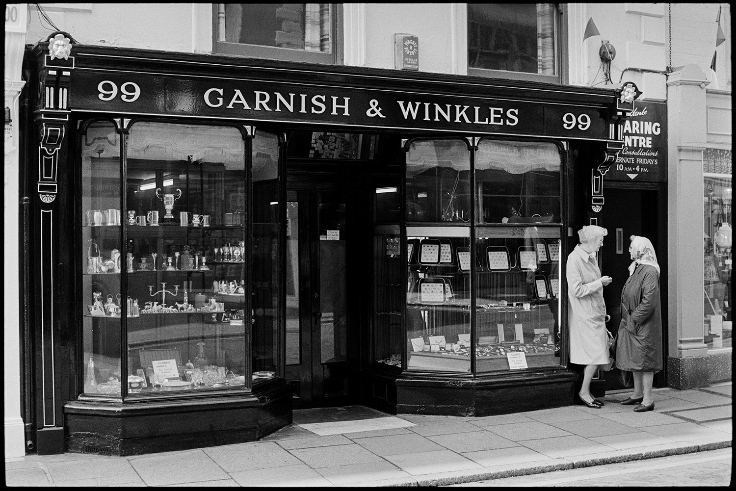 Front of jewellers shop with women chatting. 
[Two women talking outside Garnish & Winkles jewellery shop in Barnstaple High Street. Various items of jewellery and silver items are displayed in the window.]