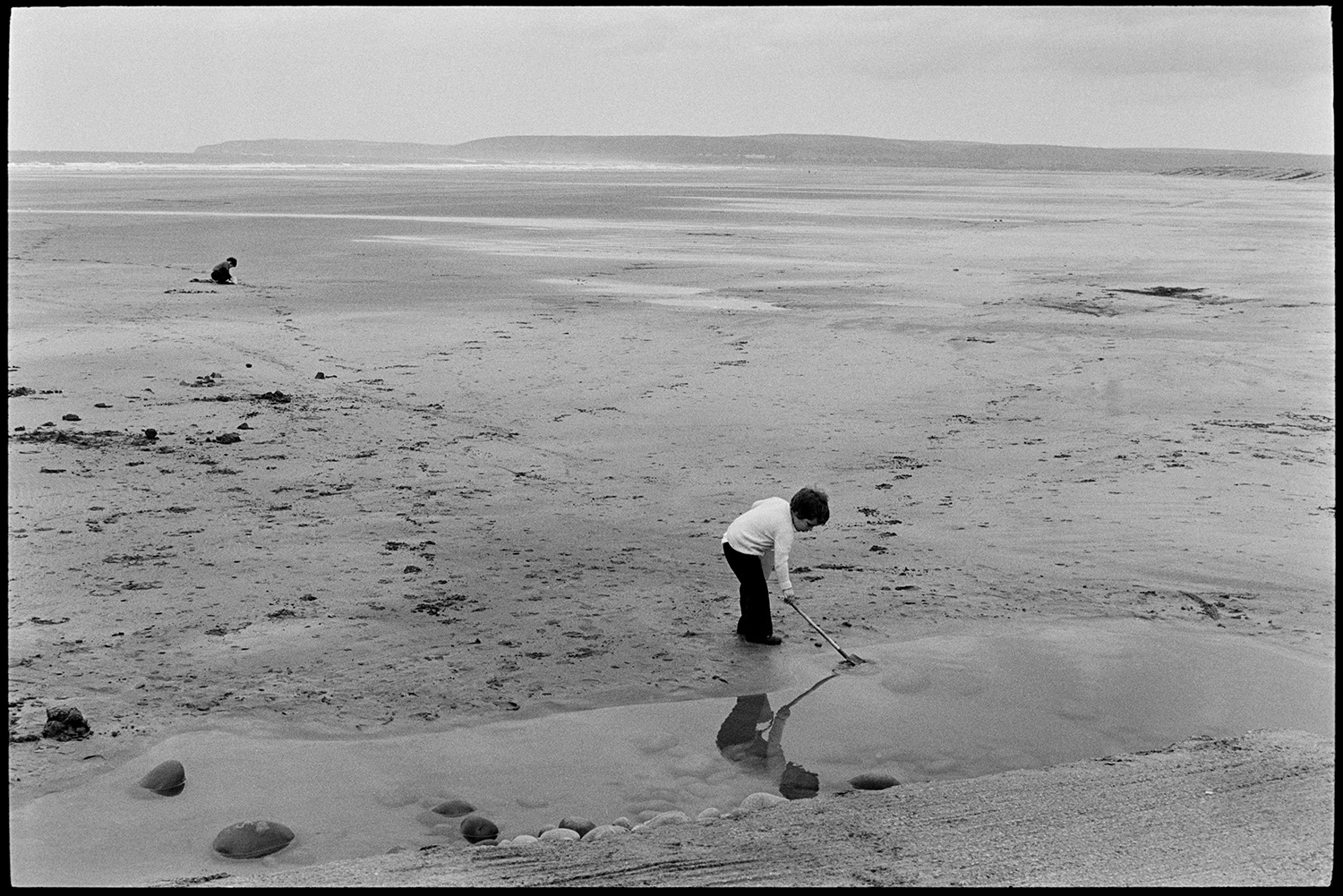 Beach scene with child and dog. 
[Two children playing on the beach at Westward Ho! One child is playing in a rock pool with a stick.]