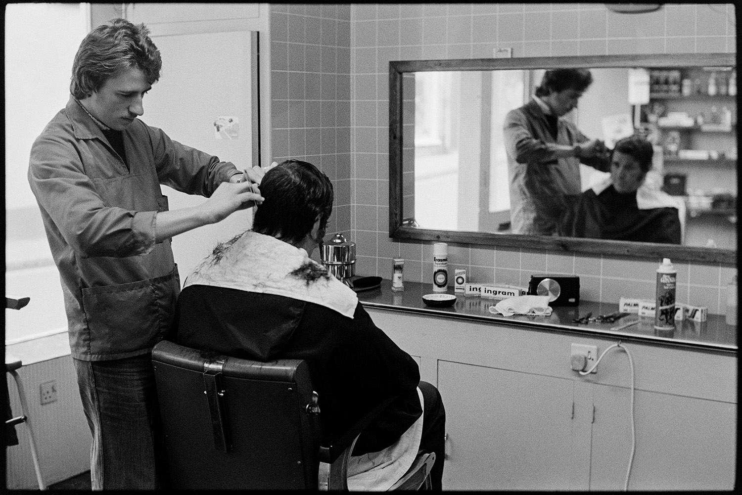 Barber cutting man's hair. 
[A barber cutting a man's hair in a barber shop in Torrington. A mirror is on the wall and various items including hairspray and scissors are on the counter.]