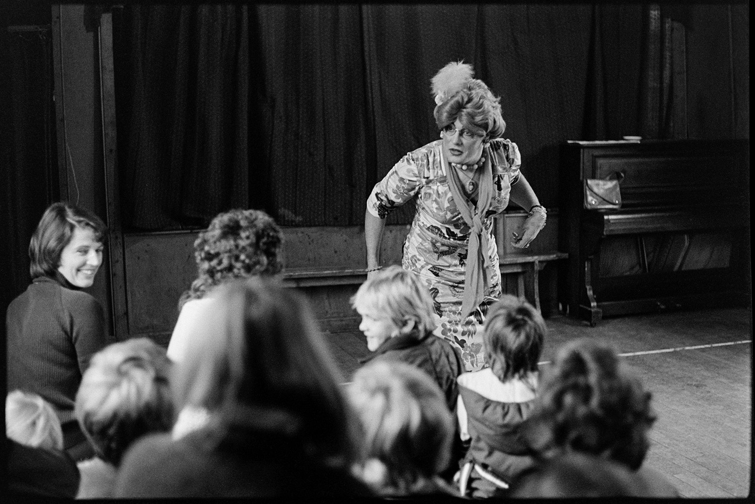 Drag artist in school, with children. 
[The drag artist, Henrietta Sluggit performing for a group of school children and teachers in a school hall. A stage and piano are visible in the background.]