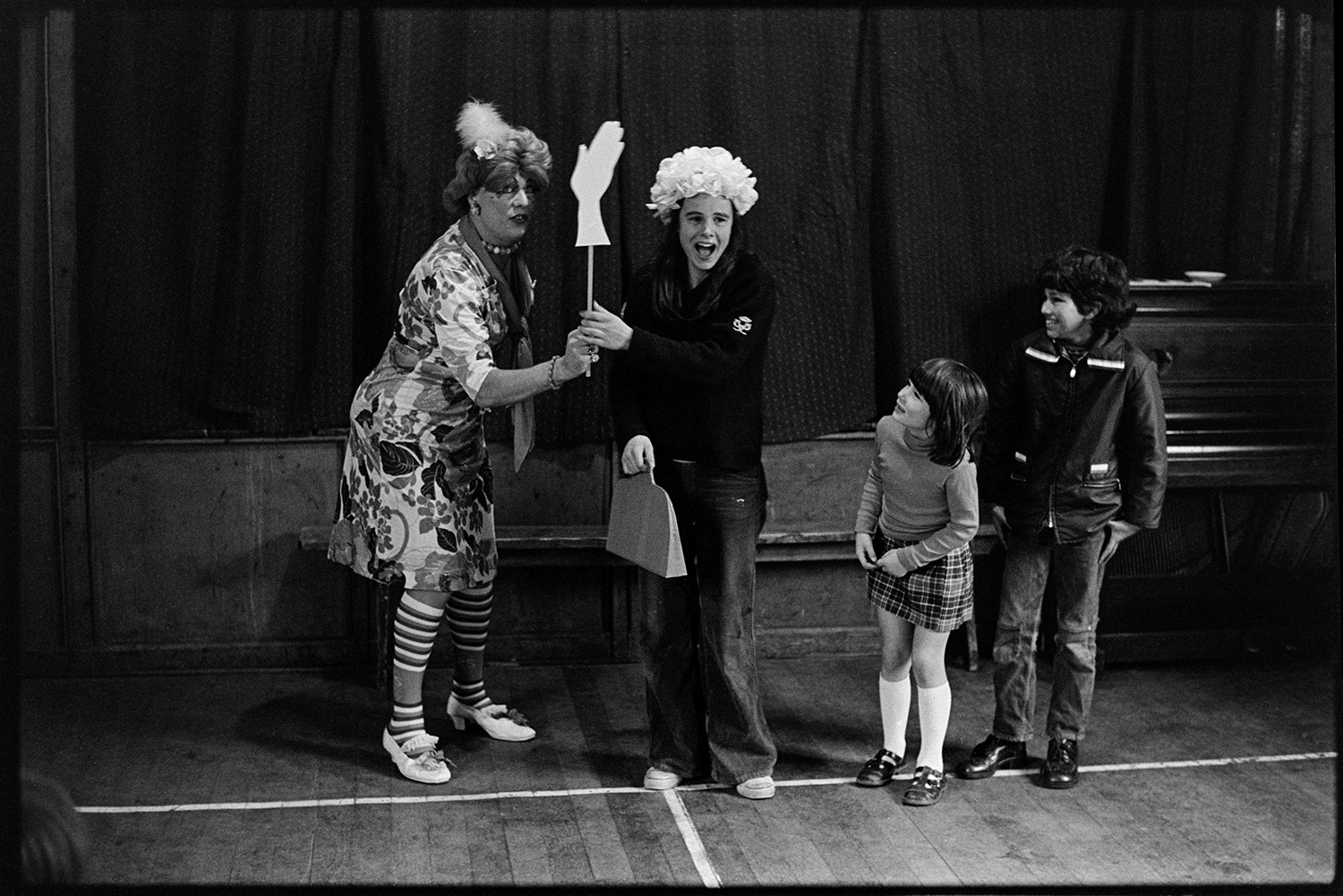 Drag artist in school, with children. 
[The drag artist Henrietta Sluggit posing with three school children in a school hall. One of the children is holding props and wearing a wig. A piano is visible in the background.]