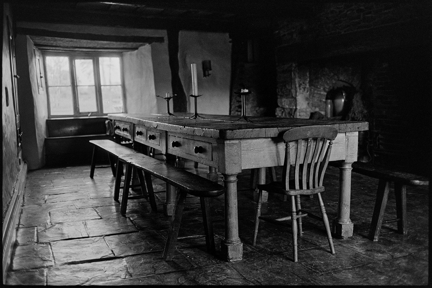 Farmhouse used as creative writing centre. 
[A large wooden dining table with drawers and benches at the Arvon Centre at Totleigh Barton, Sheepwash.  A chair is also at the table which has candlesticks placed on it. The floor is made from flagstones. The farmhouse was used as a creative writing centre.]