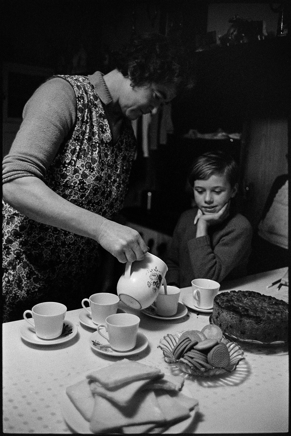 Woman making tea. Husband and children eating it. 
[Mrs Oke pouring tea for her son at Deckport, Hatherleigh. The table is laid with bread, biscuits and cake.]
