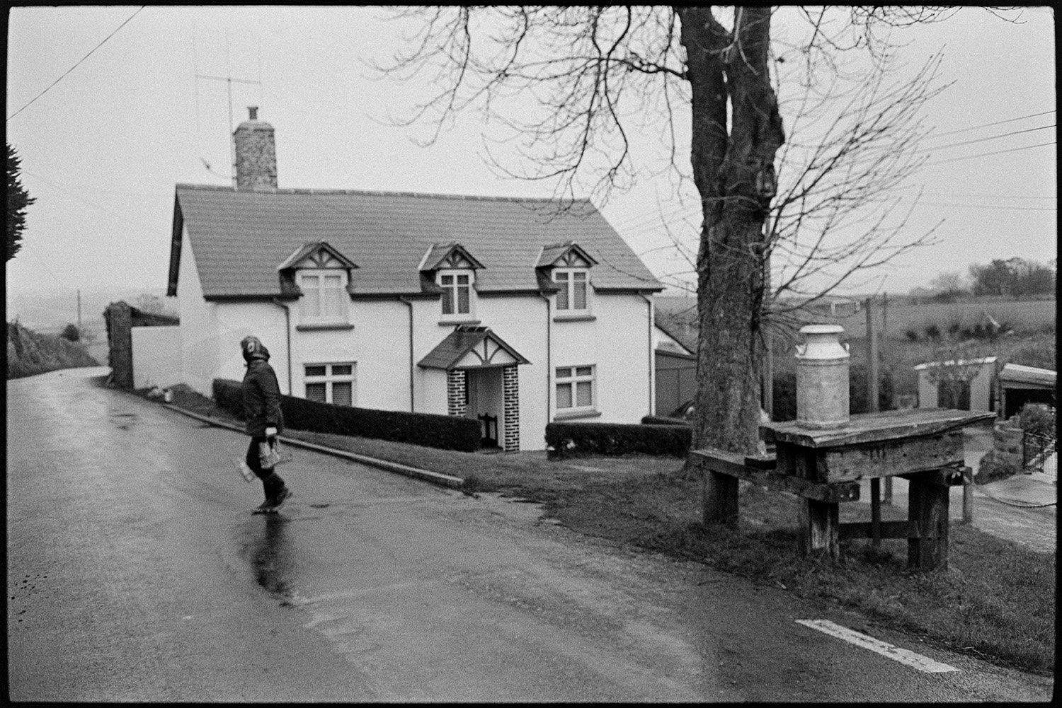 Woman delivering milk round village. <br />
[Jane Woolacott crossing a road by a house and carrying two holders containing milk bottles, on her rounds delivering milk in Atherington. A milk church is on a wooden stand under a tree in the foreground.]