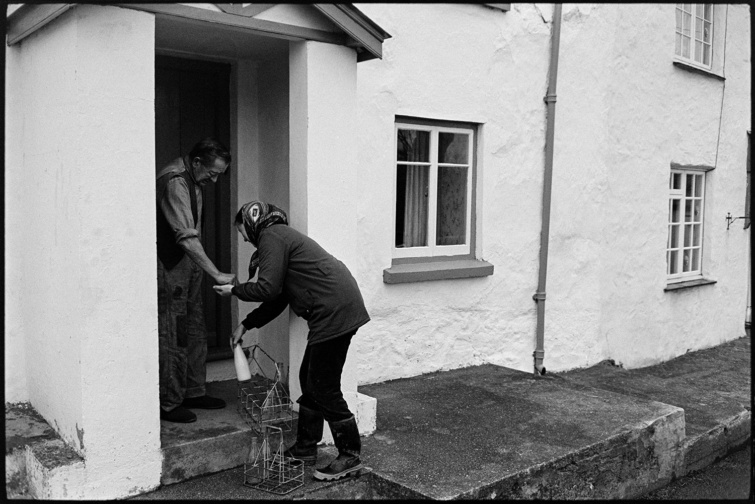 Woman delivering milk round village. <br />
[Jane Woolacott delivering and collecting milk bottles from a man at his house in Atherington. He is paying her for the milk.]