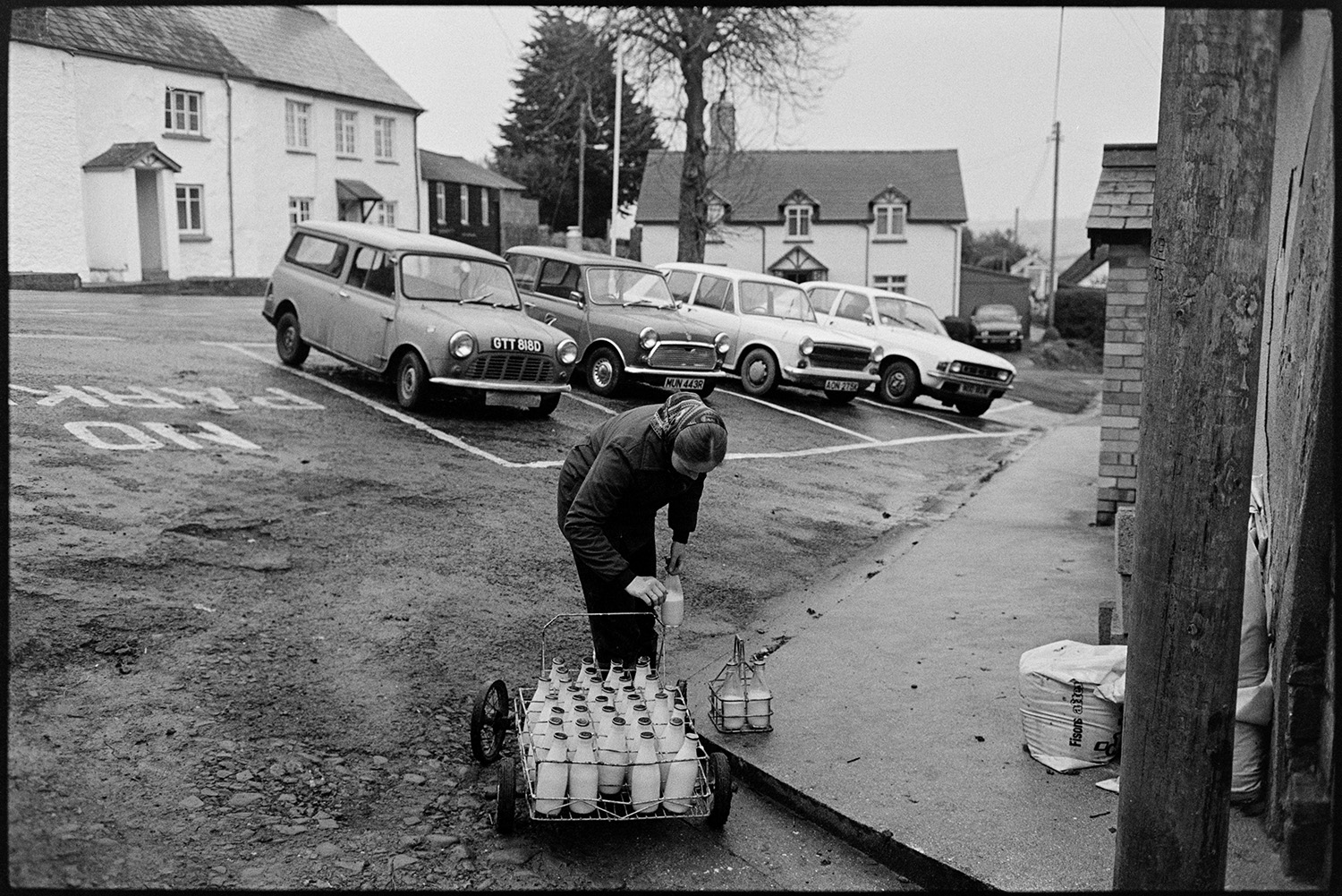 Woman delivering milk round village. <br />
[Jane Woolacott delivering milk bottles in Atherington. He is transferring bottles from a trolley into a small holder outside a house. Parked cars and houses are visible in the background.]