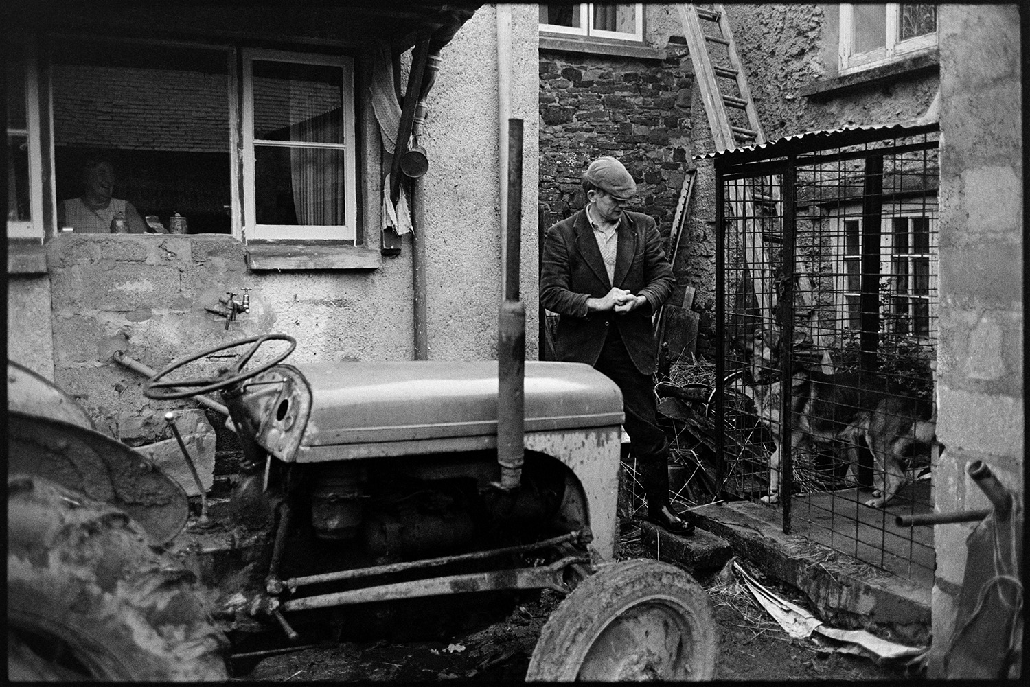 Farmer at farm door, with dog. 
[Jim Woolacott outside his farmhouse at Verdun, Atherington. His is stood next to a dog in a cage and a tractor.]