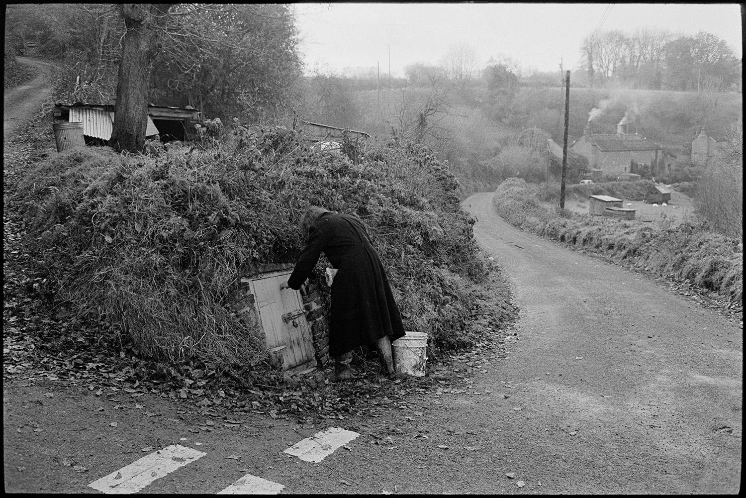 Man taking water from well. 
[Tony Webb getting water from a well behind a wooden door in a hedgebank by a road junction at Millhams, Dolton. Smoke can be seen rising from cottage chimneys in the background.]