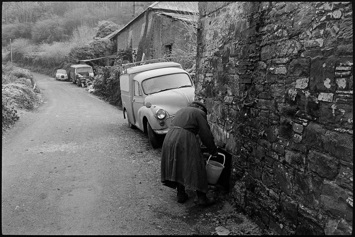 Man taking water from well at side of road. 
[Ivor Brock drawing water from a well at the side of the road and filling a bucket, at Millhams, Dolton. Vans and cars are parked further along the road.]