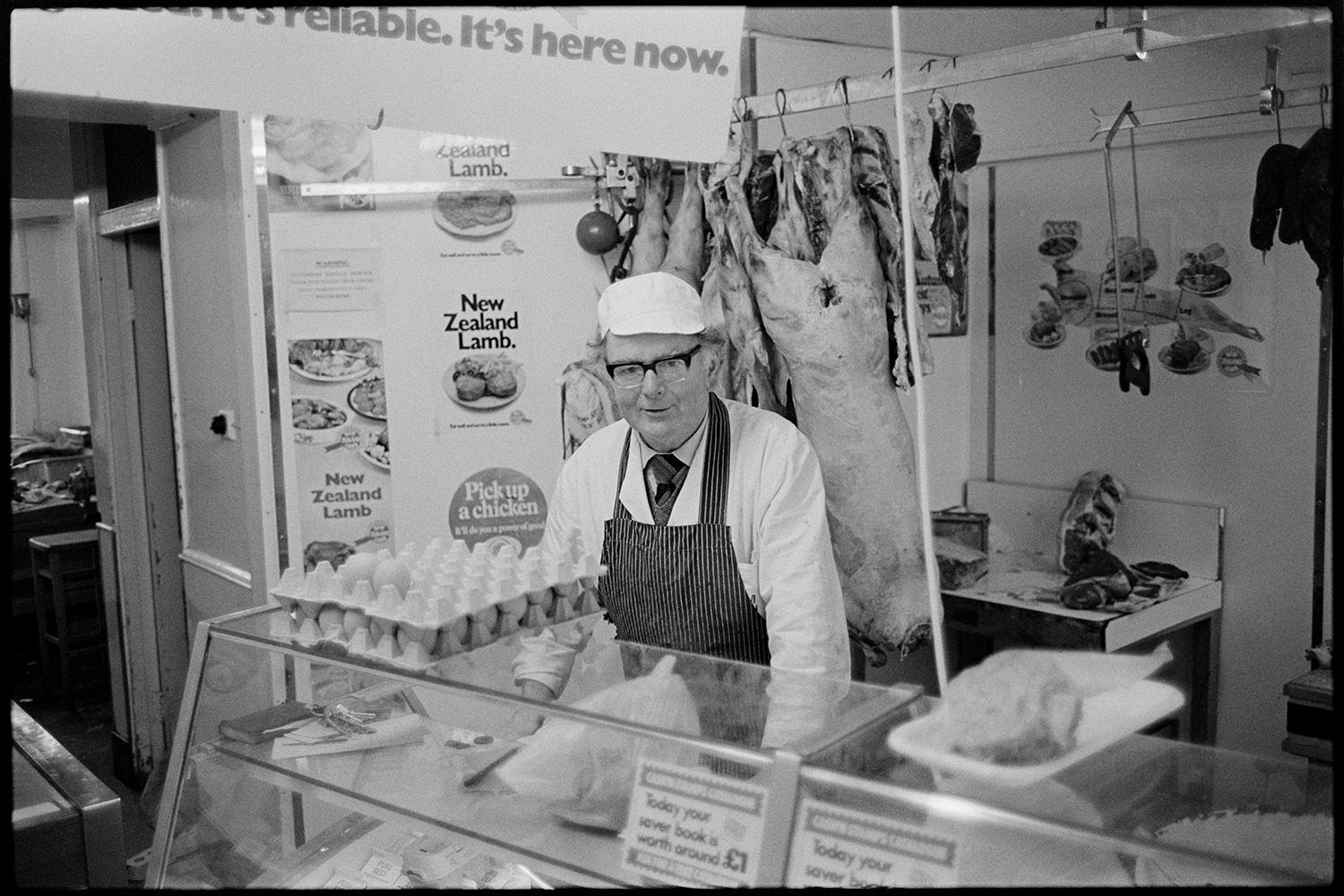 Staff of butchers shop, office with cashier. 
[A butcher stood behind the counter in a butcher's shop in Torrington. Eggs are displayed on the counter top and meat is hung up on hooks in the background.]
