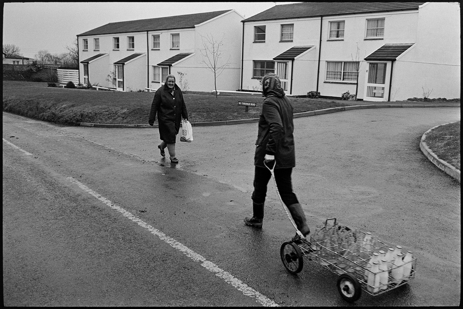 Woman delivering milk round village. <br />
[Jane Woolacott delivering milk to houses in Atherington. She is pulling a trolley with the milk bottles. Another woman is walking past her on the street.]