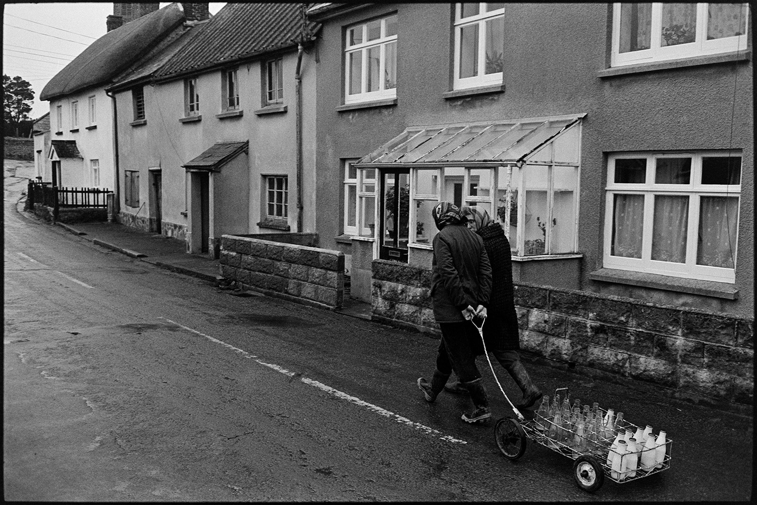 Woman delivering milk round village. <br />
[Jane Woolacott delivering milk to houses in Atherington. She is pulling a trolley with the milk bottles. Another woman is walking with her.]