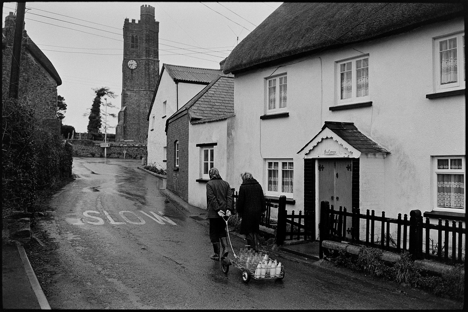 Woman delivering milk round village. <br />
[Jane Woolacott delivering milk to houses in Atherington. She is pulling a trolley with the milk bottles. Another woman is walking with her past Fern Cottage toward Atherington church tower.]