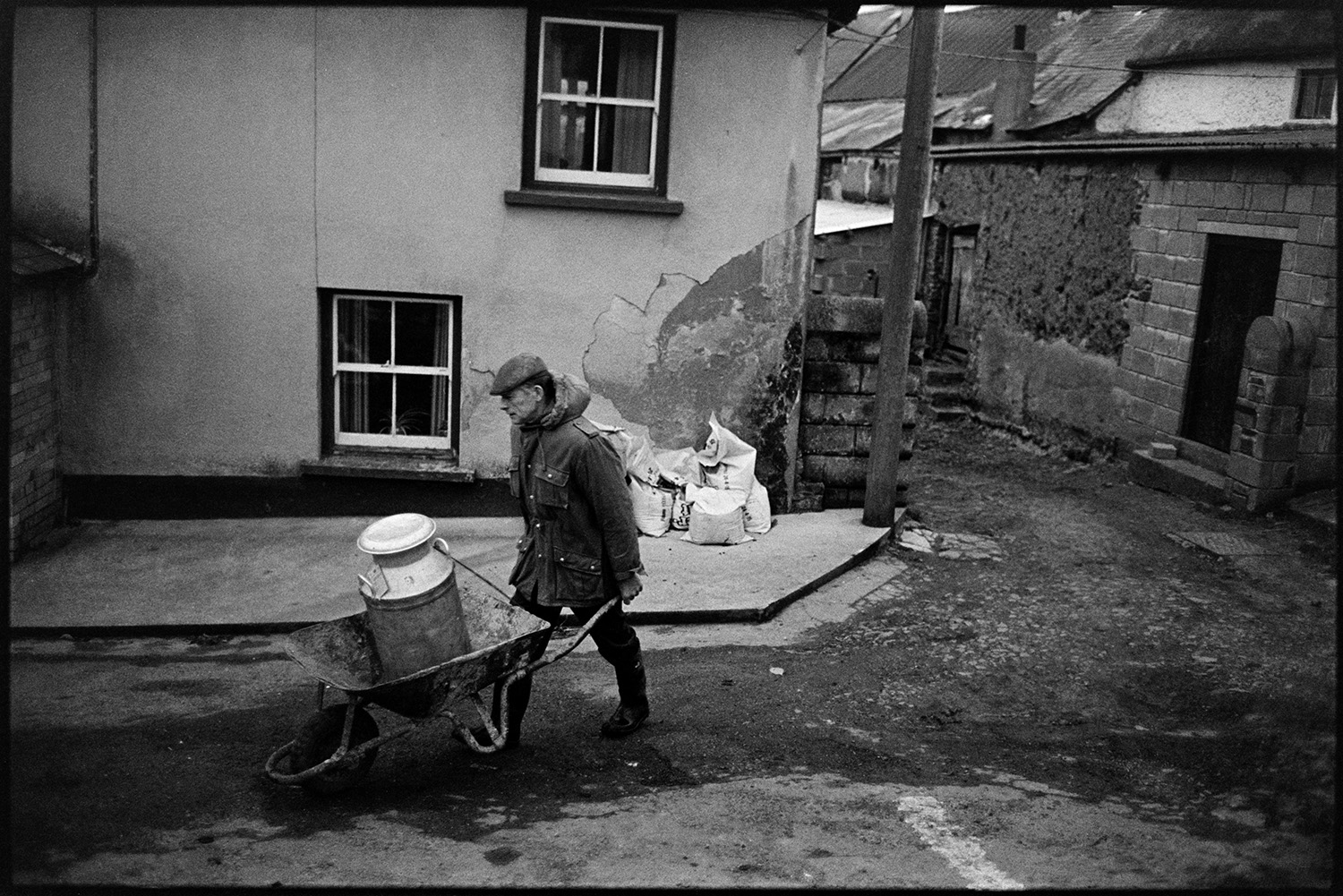 Farmer taking milk churn to stand in middle of village. 
[Jim Woolacott transporting a milk churn in a wheelbarrow from the farmyard at Verdun, Atherington to a milk churn stand in the village.]