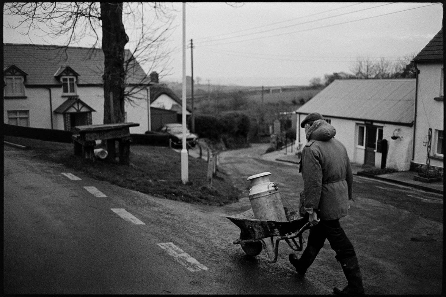 Farmer taking milk churn to stand in middle of village. 
[Jim Woolacott transporting a milk churn in a wheelbarrow to a wooden milk churn stand in Atherington.]