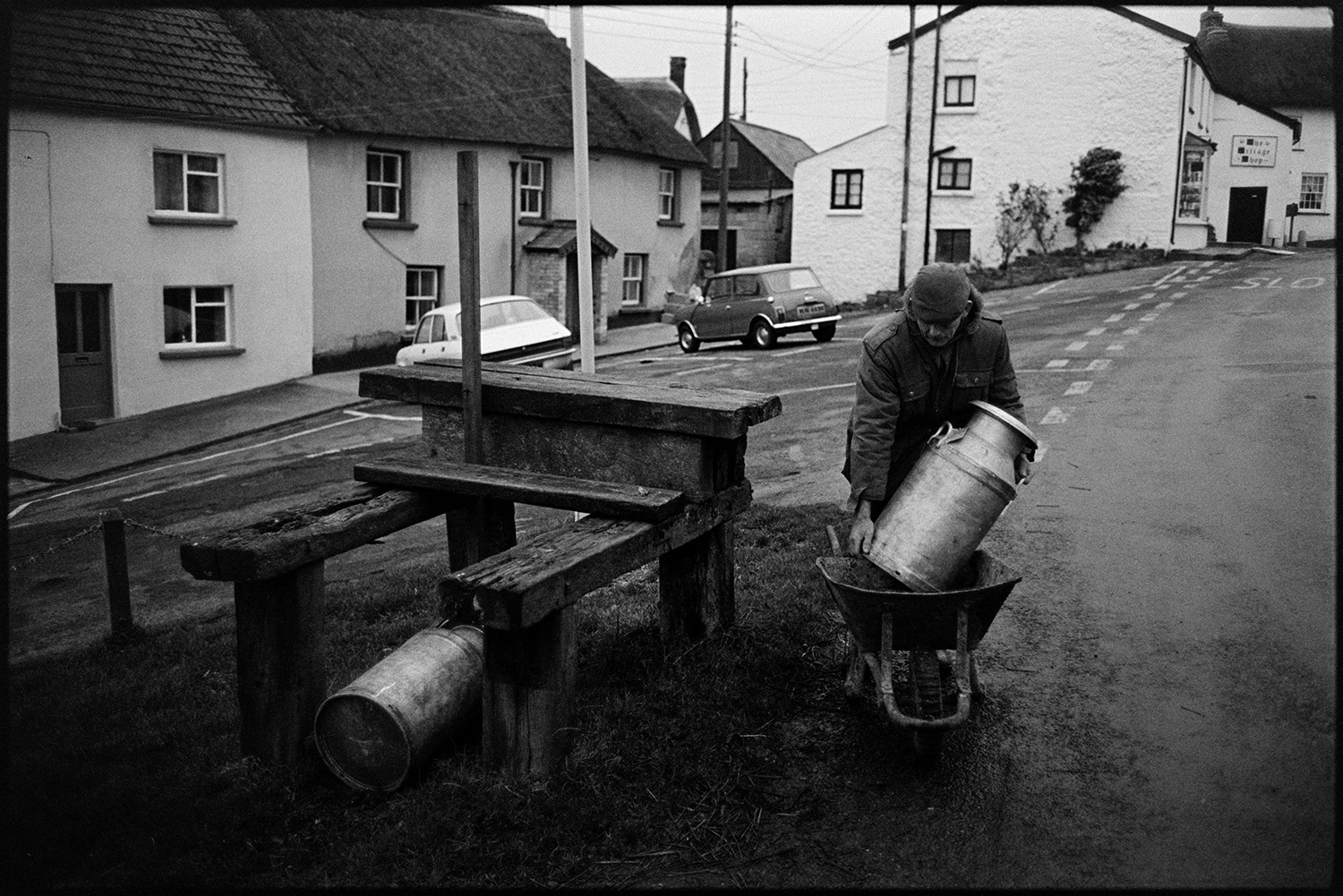 Farmer taking milk churn to stand in middle of village. 
[Jim Woolacott lifting a milk churn out of a wheelbarrow and placing it on a wooden milk churn stand in Atherington.]