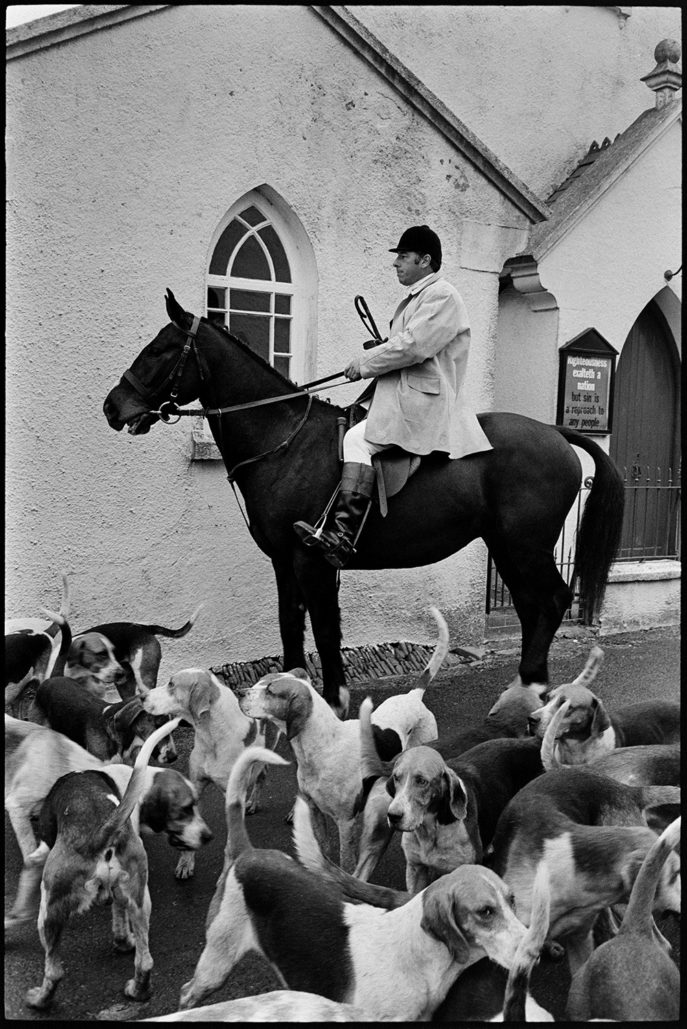 Hunt meet at village pub, with spectators, hounds and huntsmen. 
[A huntsman on horseback outside a chapel in High Bickington with hounds, before a hunt.]