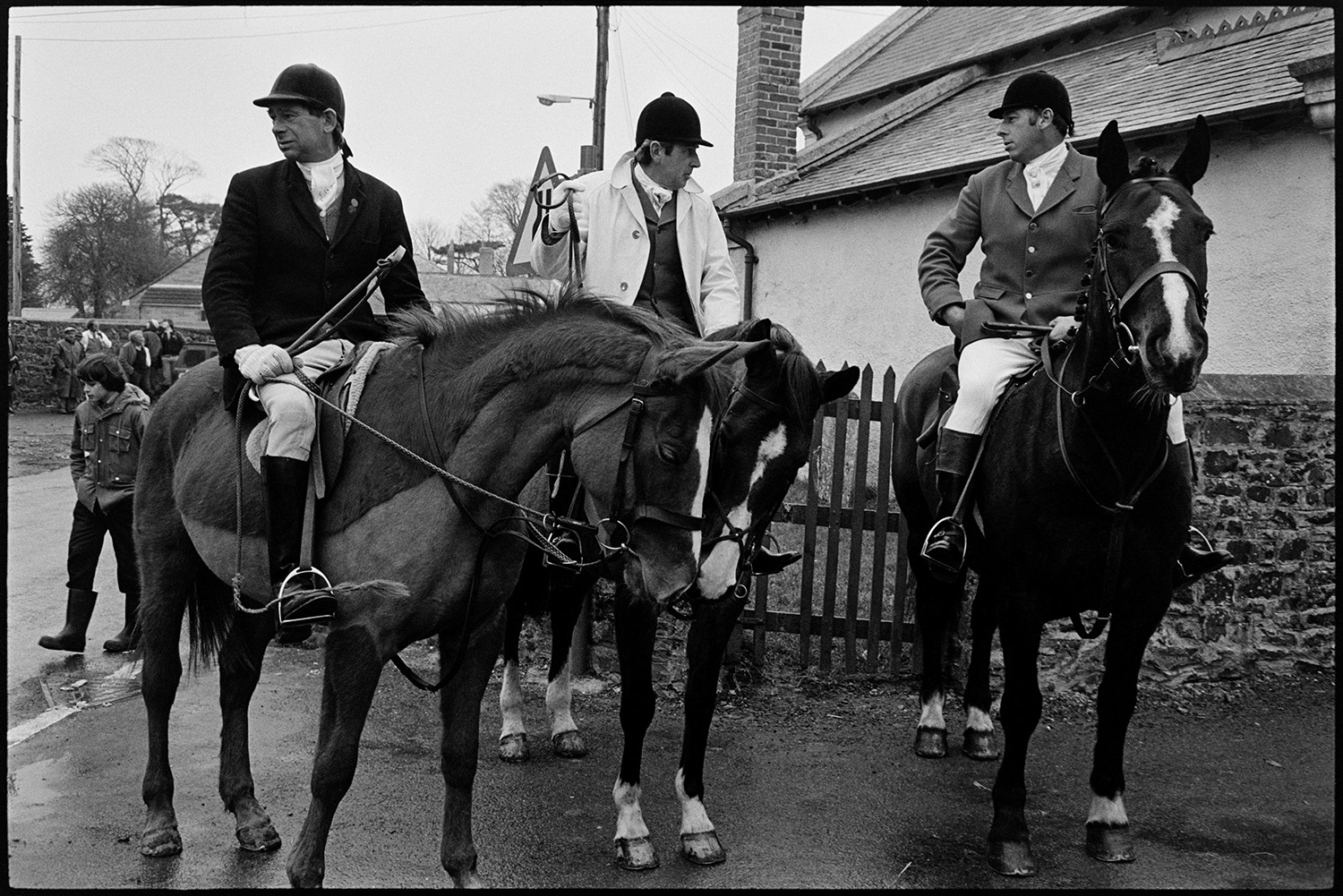 Hunt meet at village pub, with spectators, hounds and huntsmen. 
[Three huntsmen on horseback at a hunt meet in High Bickington. Spectators can be seen in the background.]