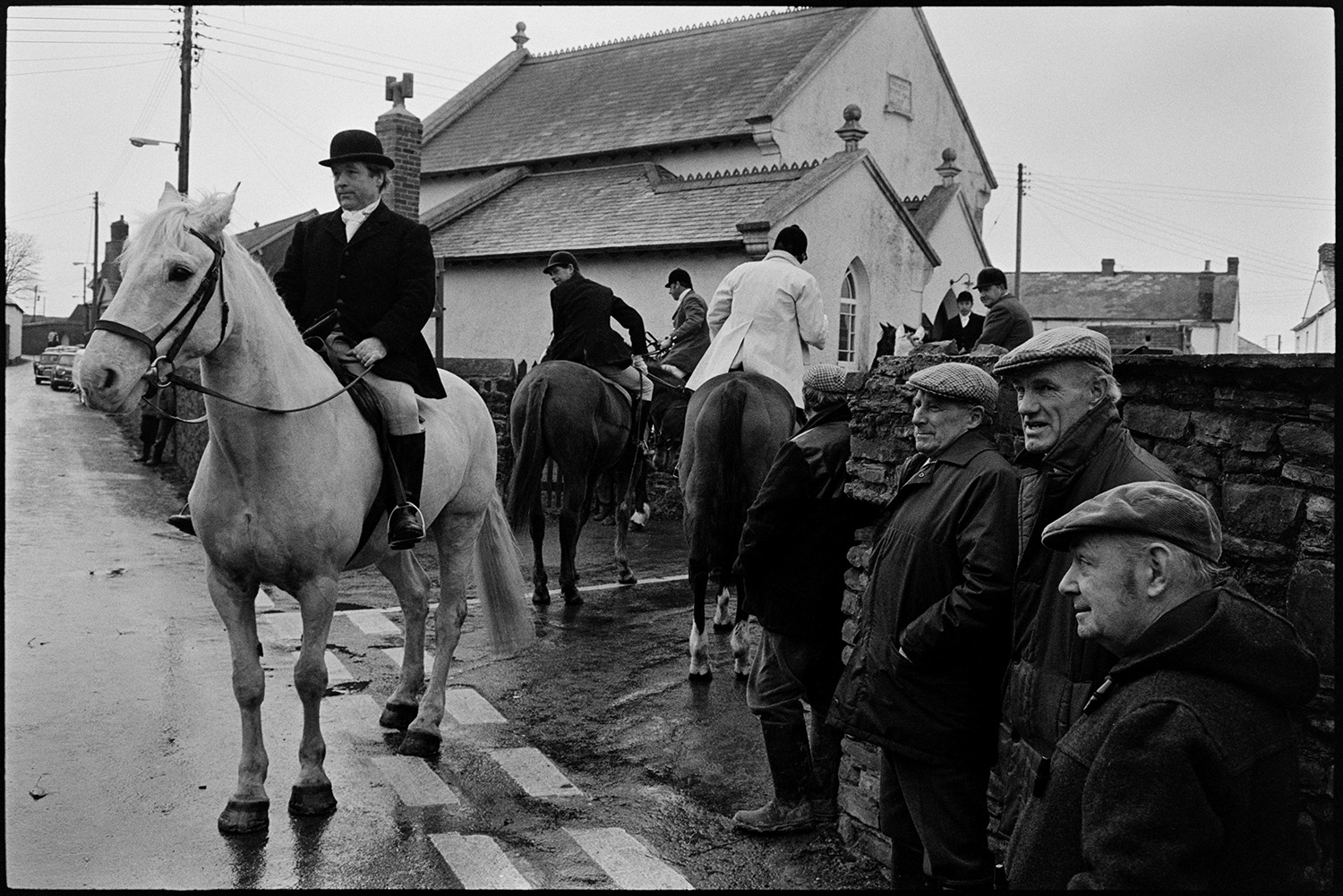 Hunt meet at village pub, with spectators, hounds and huntsmen. 
[Spectators watching mounted huntsmen at a hunt meet in High Bickington. A chapel building can be seen in the background.]