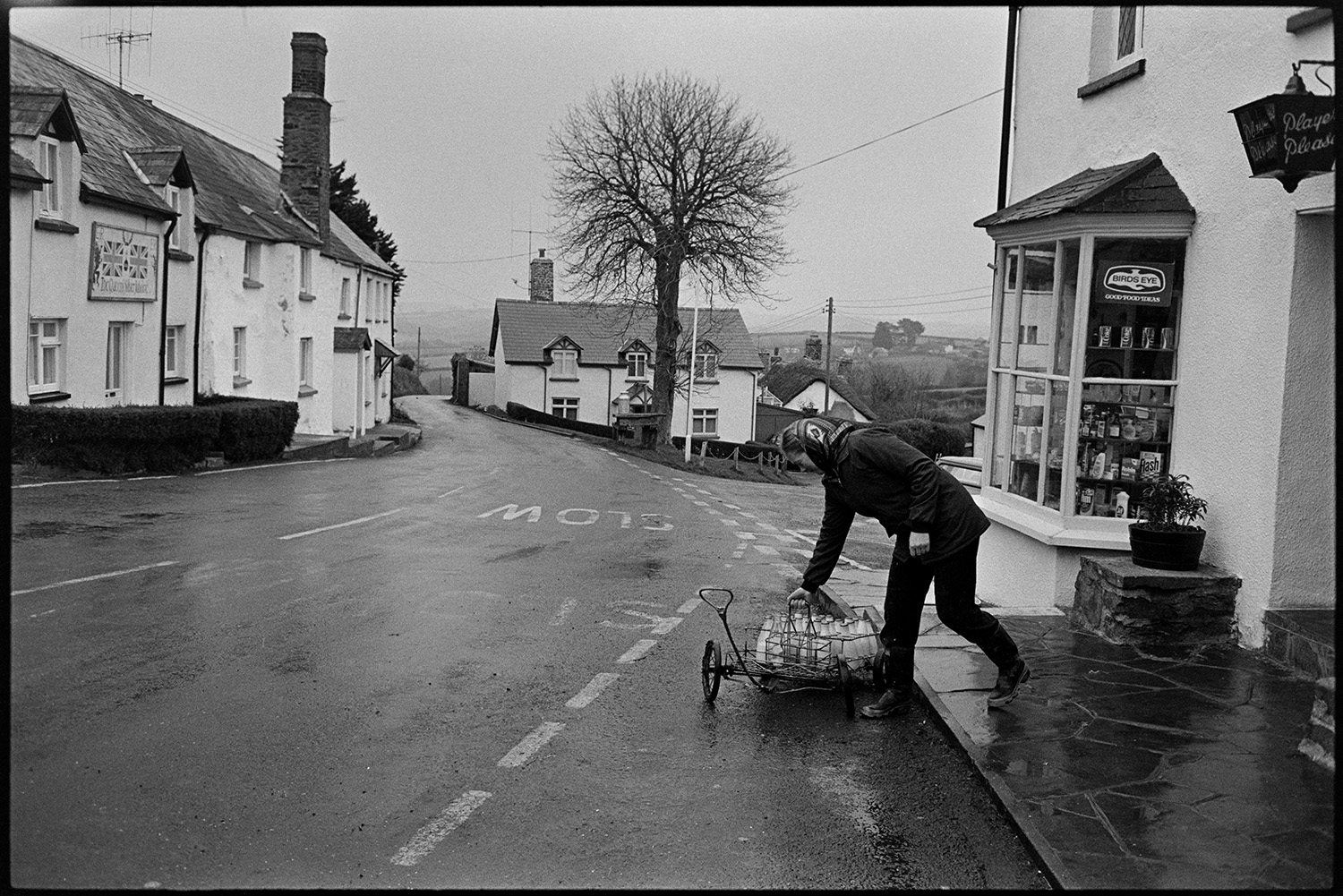 Woman doing milk round in village, chatting to customers. <br />
[Jane Woolacott delivering milk to a shop in Atherington. She is transporting the milk bottles in a trolley. On the opposite side of the road a house has a decoration up for the Silver Jubilee of Queen Elizabeth II.]