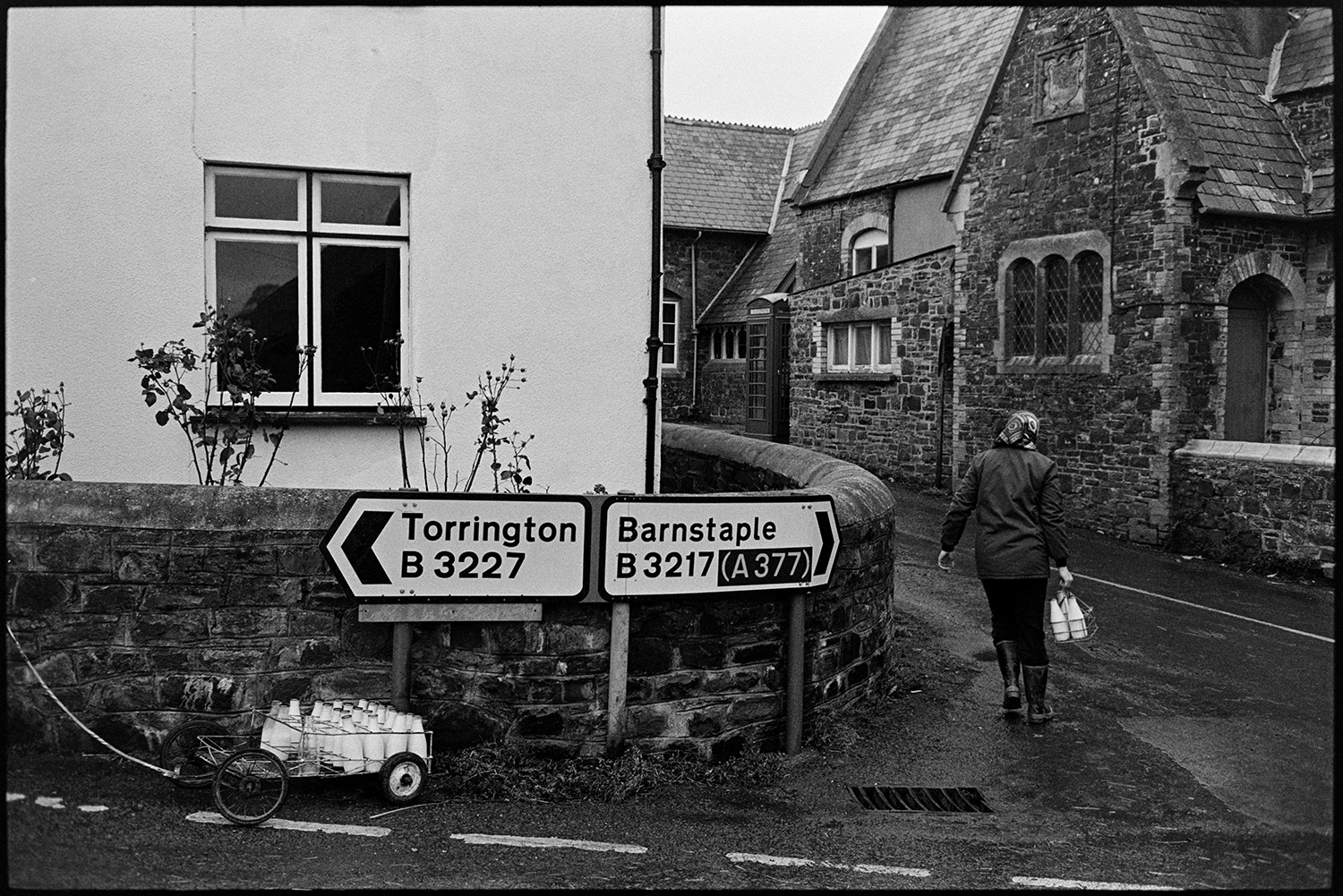 Woman doing milk round in village, chatting to customers. <br />
[Jane Woolacott delivering milk to houses in Atherington. She is transporting the milk bottles on  trolley which is parked by road signs.]