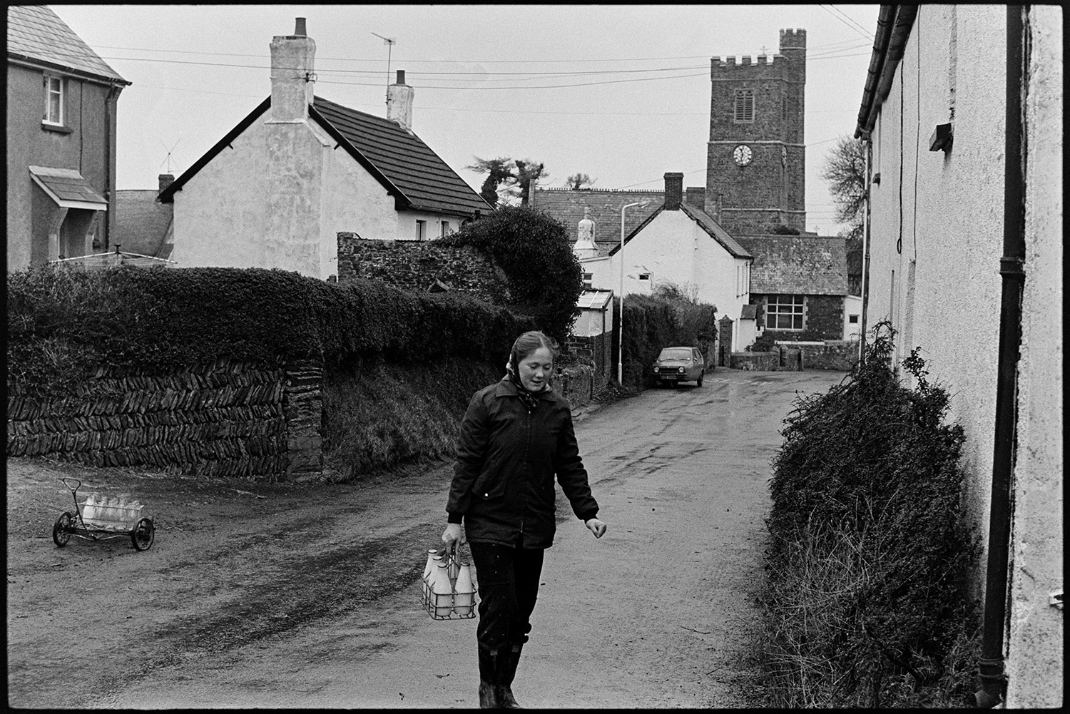 Woman doing milk round in village, chatting to customers. <br />
[Jane Woolacott delivering milk to houses in Atherington. She is transporting the milk bottles using a trolley. The church tower is visible in the background.]