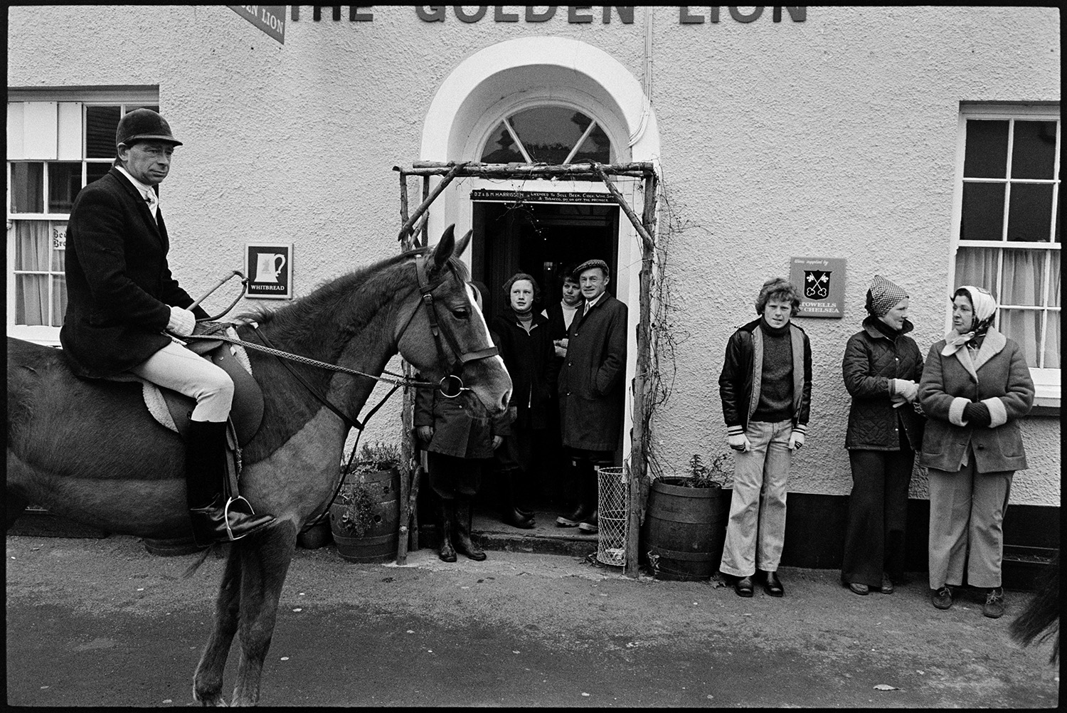 Hunt meet at village pub, with spectators, hounds and huntsmen. 
[Spectators watching a huntsman on horseback at a hunt meet outside The Golden Lion pub in High Bickington.]