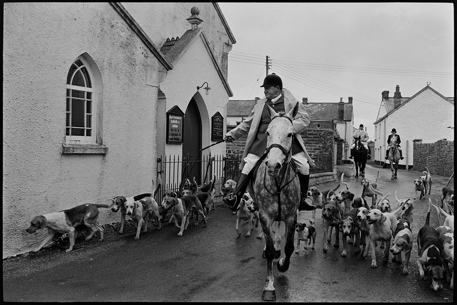 Hunt meet at village pub, with spectators, hounds and huntsmen. 
[Les Grills on horseback leading hounds and other huntsmen on horseback past a chapel in High Bickington before a hunt.]