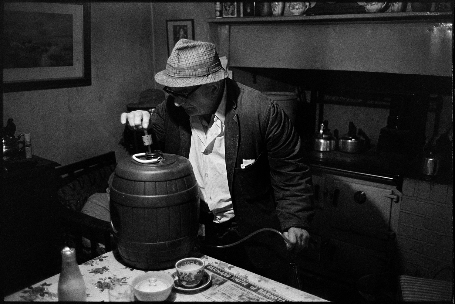 Man making home brewed beer. 
[Archie Parkhouse making beer in his kitchen at Millhams, Dolton. He is looking into the barrel of beer. A rayburn can be seen under a mantelpiece in the background.]