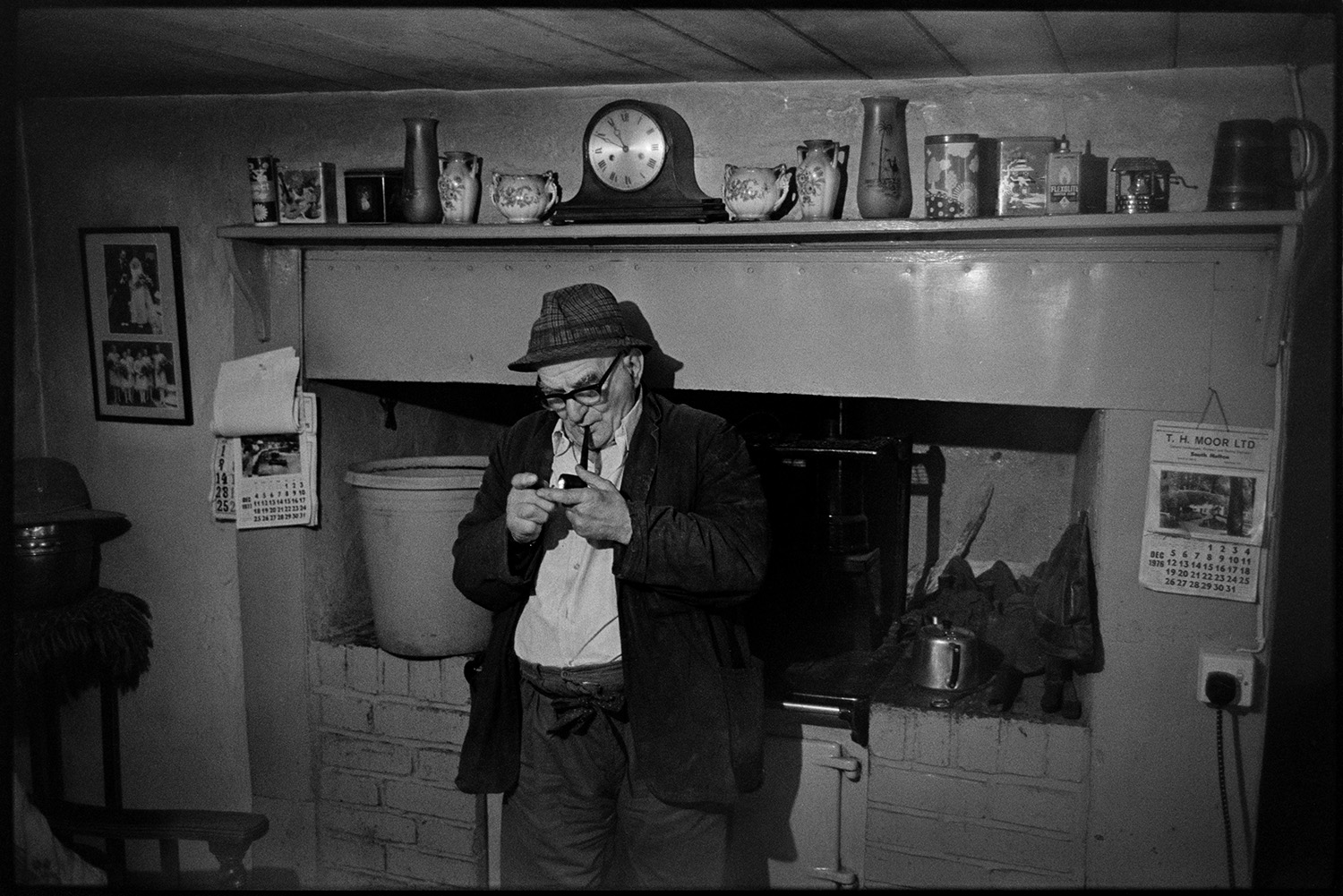 Man lighting pipe in front of mantelpiece. 
[Archie Parkhouse lighting his pipe in his kitchen at Millhams, Dolton. A rayburn stove and mantelpiece can be seen in the background. A clock and china vases are displayed on the mantelpiece.]