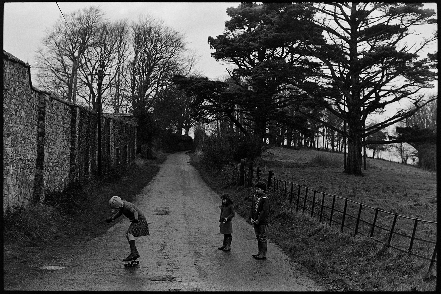Children skateboarding. 
[A girl skateboarding along a road by a stone wall at Halsdon, Dolton. Two other children are watching.]