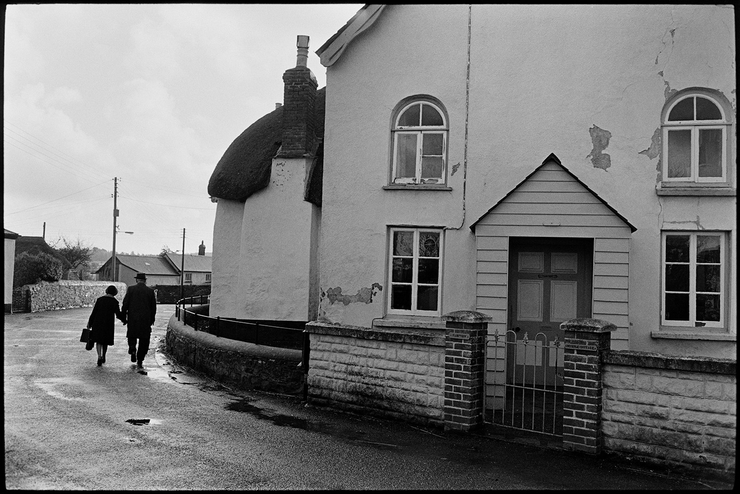 Couple walking past house with porch. 
[A man and woman walking hand in hand along West Lane, Dolton. They are passing a thatched cottage and a house with a porch.]