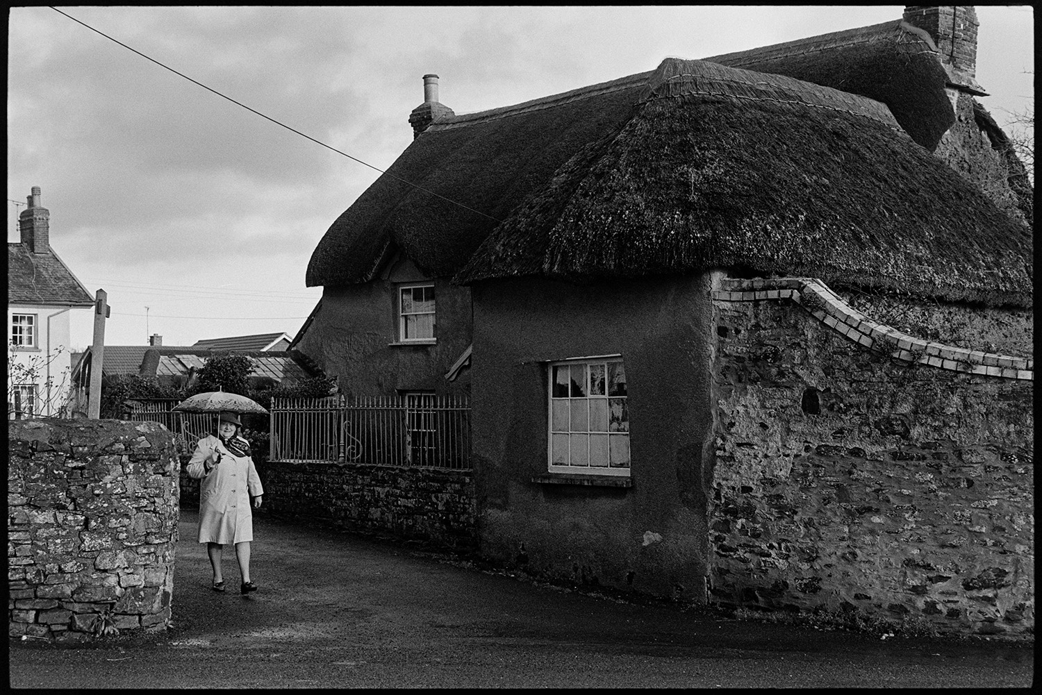 Couple walking past house with porch. 
[A woman walking along West Lane in Dolton holding an umbrella. She is passing a thatched cottage with railings outside.]