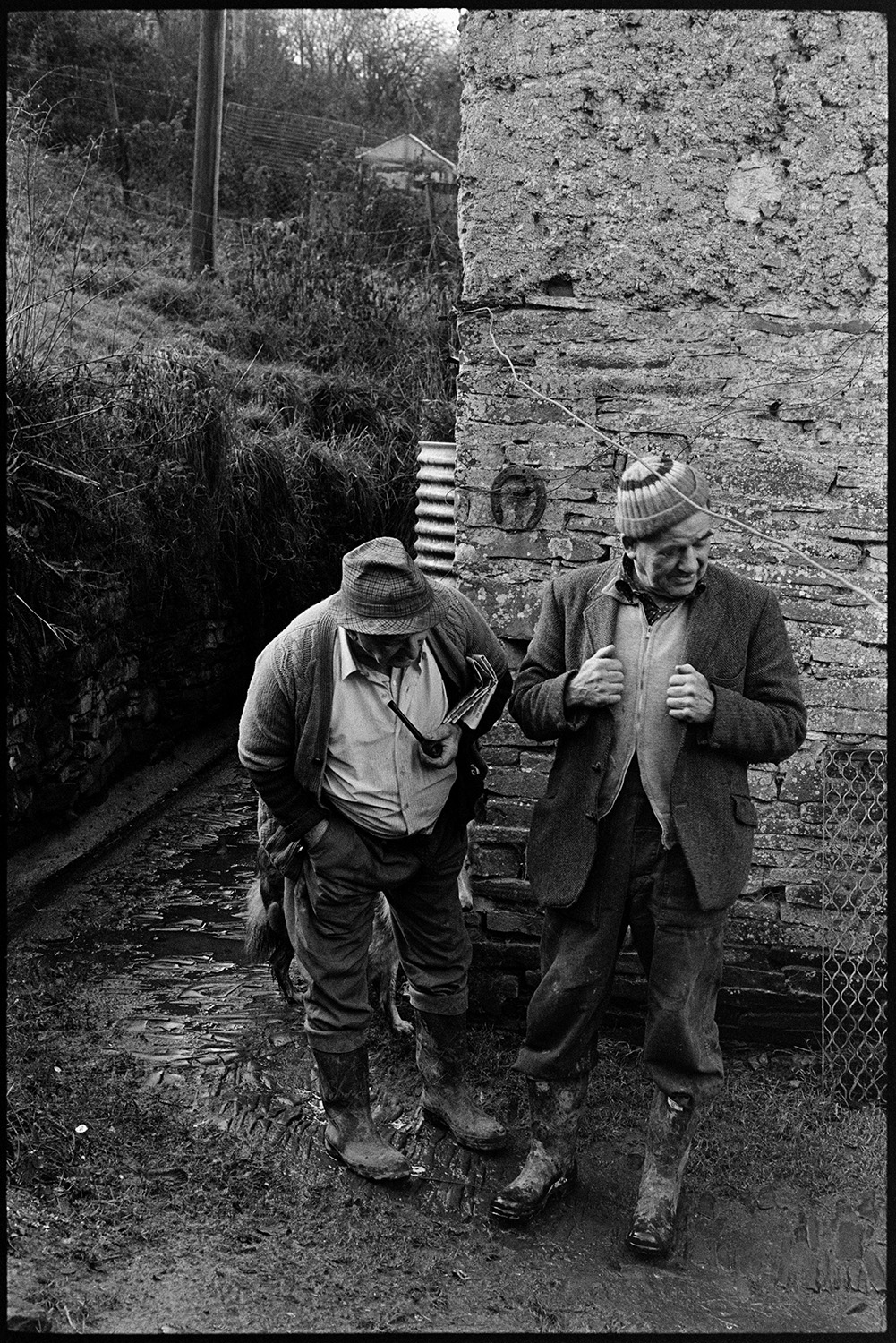 Two farmers chatting. 
[Archie Parkhouse, holding a pipe, and Frank Pickard chatting by a stone wall at Millhams, Dolton. A horse show is hung on the wall behind them. They are both wearing hats and wellington boots.]