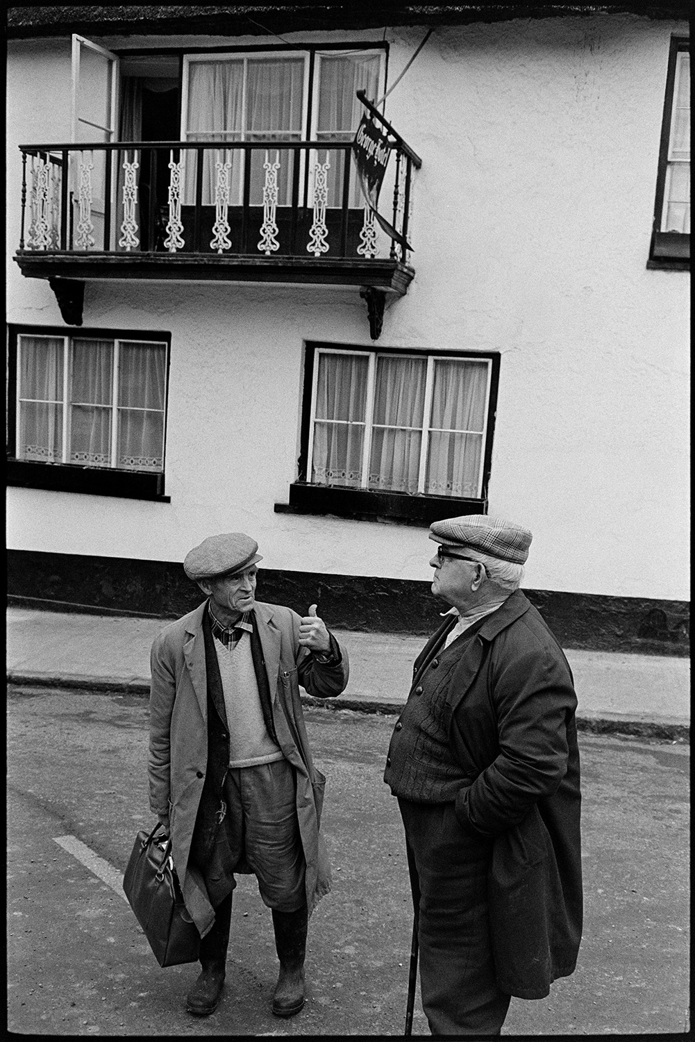 Two men chatting in front of pub. 
[Two men talking in front of the George Hotel in Hatherleigh. A balcony on the hotel is visible in the background.]