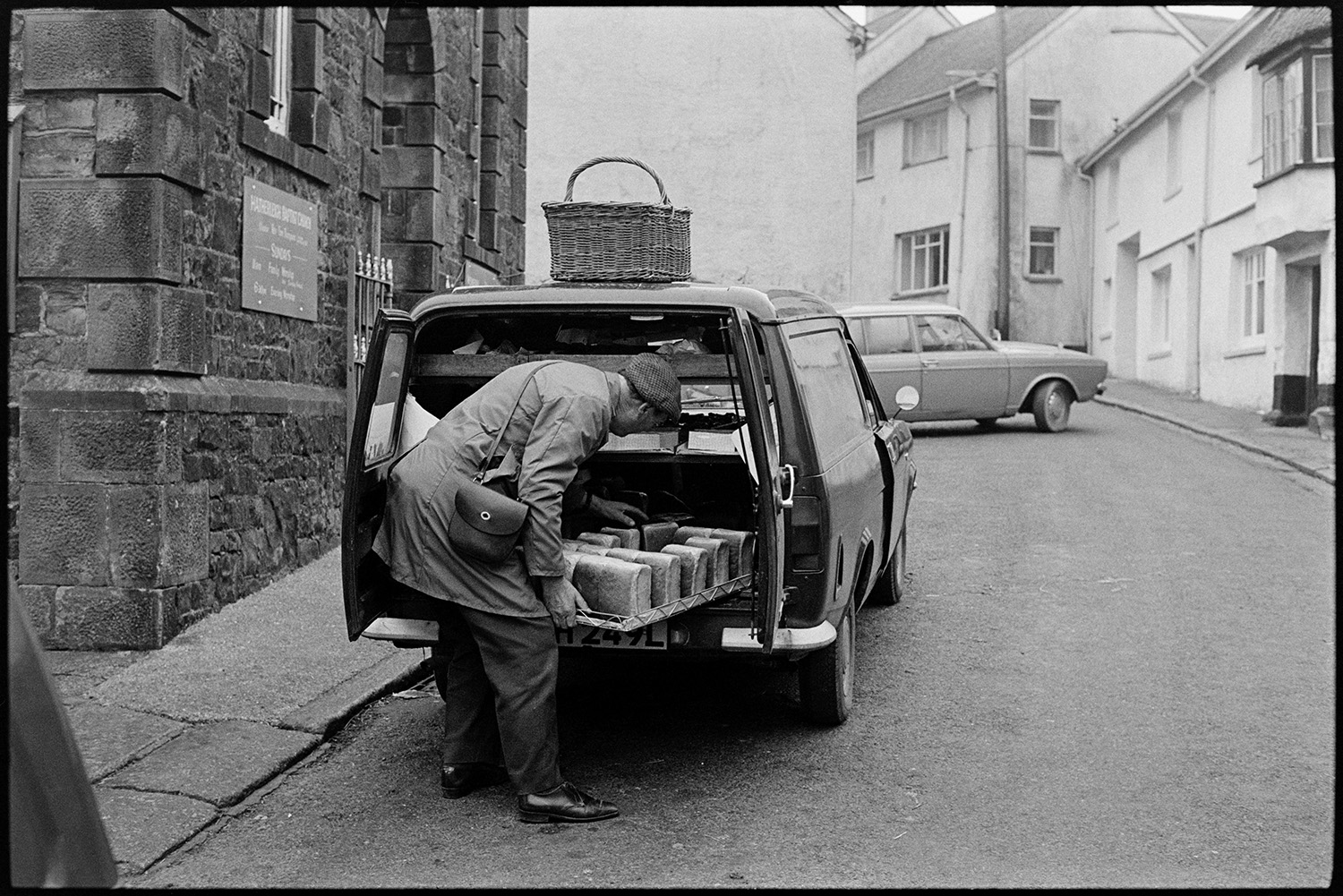 Baker selling bread from van. 
[A man unloading a tray of bread from his baker's van in a street in Hatherleigh. A basket is on the roof of the van.]