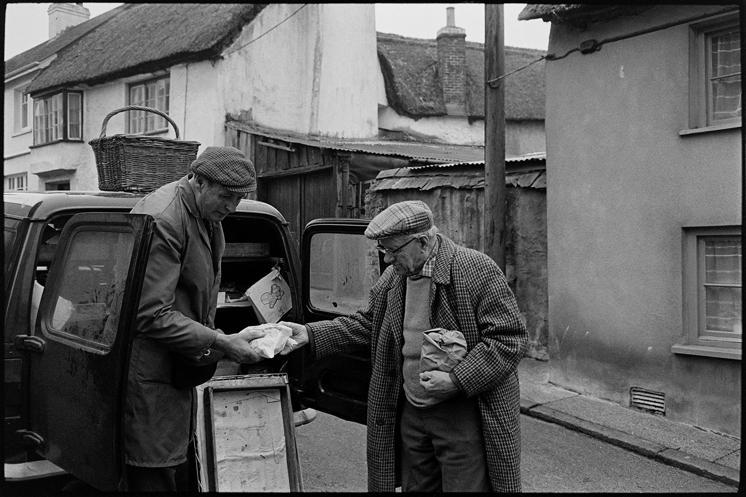 Baker selling bread from van. 
[A man buying bread from a baker's van in a street in Hatherleigh. A basket is on top of the van.]
