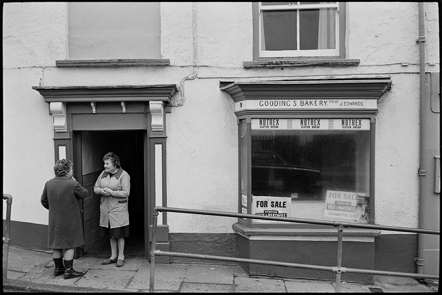 Women chatting at door of baker's shop. 
[Two women talking outside the shop front of Gooding's Bakery in Market Street, Hatherleigh. For Sale signs are up in the bakery window.]