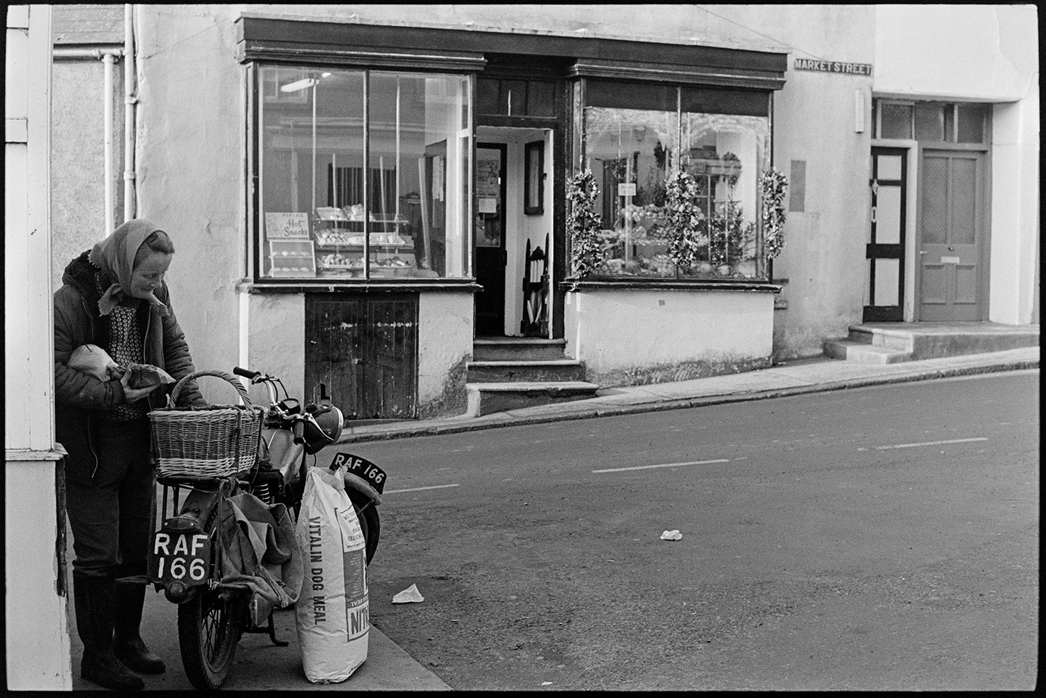 Woman motorcyclist in front of shop, street scenes with woman in telephone kiosk. 
[A woman putting groceries into a basket on a motorbike, parked opposite a shop front on Market Street in Hatherleigh. A bag of dog food is next to the motorbike.]